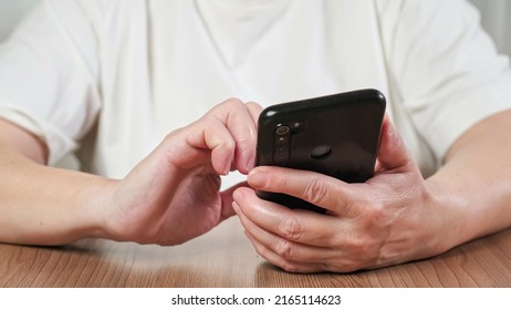 Elderly Woman In White T-shirt Learns To Use Modern Mobile Phone. Woman Scrolls Pages Of Social Media On New Black Smartphone Closeup