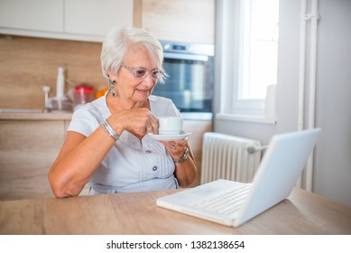 Elderly woman working on laptop computer, smiling, drinking tea.  Senior woman using laptop. Elderly woman sitting at home, using laptop computer, smiling. 