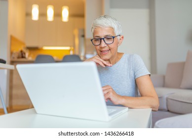 Elderly Woman Working On Laptop Computer, Smiling. Woman Working From Home On Laptop In Modern Apartment. Trendy Woman Working On Laptop From Home