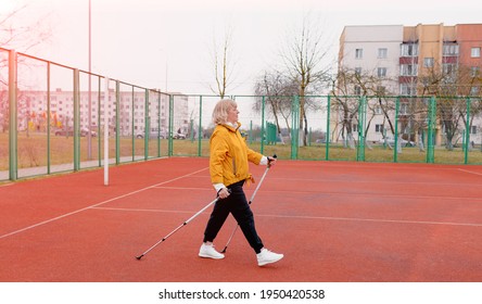 An Elderly Woman In A Yellow Sports Jacket Practices Nordic Walking Outdoors On The Stadium's Rubber Treadmill. A Sunny Sunset. Older Women Walk With A Nordic Gait, Using Trekking Poles.