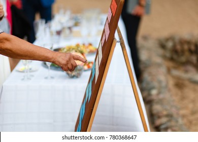 The Elderly Woman's Hand Points To The Chalkboard Seating Chart. Wedding Seating Chart On The Easel At Light Restaurant On Fresh Air.