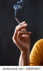 Elderly Wrinkled Human  With A Cigarette In Hand. Adult Woman Holds Cigarette With Smoke In Her Hand On A Black Background. Concepts Of Women's Health, Bad Habits. Shallow Depth Of Field