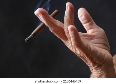 Elderly Wrinkled Human With A Cigarette In Hand. Adult Woman Holds Cigarette With Smoke In Her Hand On A Black Background. Concepts Of Women's Health, Bad Habits. Shallow Depth Of Field