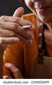 Elderly Wrinkled Woman With A Cigarette In Hand.  Older, Adult Woman Holds Cigarette In Her Hand On Table. Concepts Of Women's Health, Bad Habits, Addiction. Shallow Depth Of Field