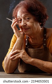 Elderly Wrinkled Woman With A Cigarette In Hand.  Older, Adult Woman Holds Cigarette In Her Hand On Table. Concepts Of Women's Health, Bad Habits, Addiction. Shallow Depth Of Field