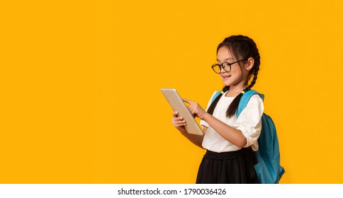 E-Learning. Asian School Girl Using Digital Tablet Doing Homework Online Standing Over Yellow Studio Background. Panorama With Empty Space