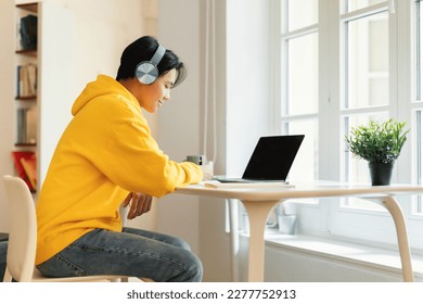 E-Learning. Asian Teenager Guy Sitting At Laptop Doing Homework Wearing Earphones And Taking Notes Sitting At Table At Home. Smiling Student Writing Essay Studying Online. Side View - Powered by Shutterstock