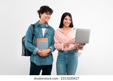 E-Learning. Cheerful Students Couple Using Laptop Computer For Educational Purposes Standing With Backpack And Workbooks Over White Studio Background. Friends Duo Studying Online Together