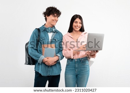 E-Learning. Cheerful Students Couple Using Laptop Computer For Educational Purposes Standing With Backpack And Workbooks Over White Studio Background. Friends Duo Studying Online Together