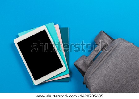 E-learning concept - blank tablet computer on top of stack of colorful books and notebooks and backpack on blue background