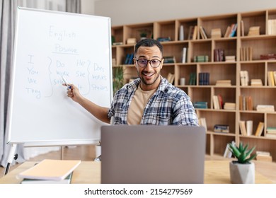 E-learning concept. Excited male English teacher sitting at table, pointing with pen at whiteboard, explaining grammar rules to students, having online lesson from home - Shutterstock ID 2154279569