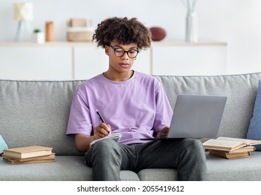 E-learning Concept. Focused African American Teenager Having Remote Lesson On Laptop, Taking Notes, Sitting On Sofa At Home. Serious Black Teen Participating In Online Conference Or Lecture