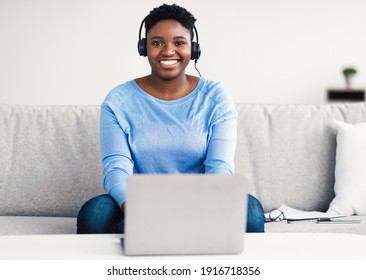 E-learning Concept. Happy Black Woman Wearing Headset Studying Online With Laptop At Home, Cheerful Casual African American Lady Sitting On Sofa At Desk With Computer, Looking At Camera