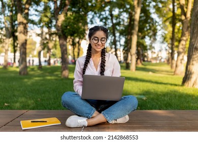 E-learning concept. Indian female student with laptop sitting on bench and watching educational webinar, wearing eyeglasses and smiling. Distance college education - Shutterstock ID 2142868553