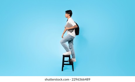 E-Learning Concept. Japanese Teen Guy Holding Laptop Computer Walking Upstairs On Ladder Posing On Blue Studio Background. Online Education Concept. Full Length, Panorama