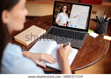 E-learning Concept. Over the shoulder back view of female student sitting at table, writing in notebook, taking notes, using laptop, having online class with English teacher. Distant studying