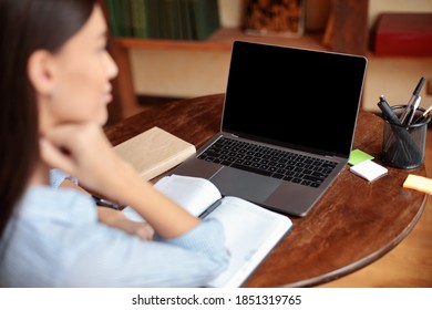 E-learning Concept. Over The Shoulder View Of Woman Sitting At Desk With Open Notebook, Using Laptop With Black Empty Screen For Mockup Template, Watching Webinar, Studying Online At Home