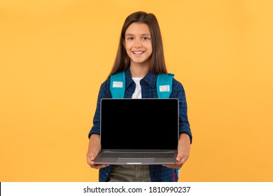 Elearning Concept. Portrait Of Beautiful Girl Holding And Presenting Laptop With Black Screen For Mock Up, Orange Wall