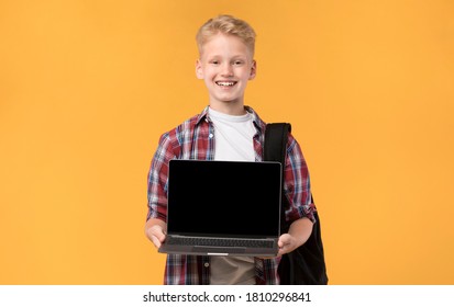 Elearning Concept. Portrait Of Blond Boy Holding And Presenting Laptop With Black Screen For Mock Up, Orange Wall