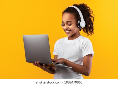 E-learning Concept. Portrait of smiling African American teenage girl holding and using laptop over yellow orange studio background, wearing wireless headphones, typing on keyboard