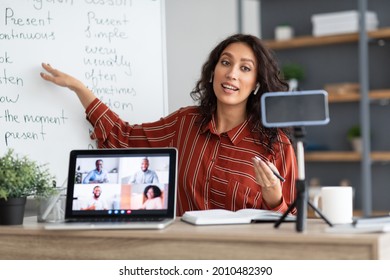 E-learning Concept. Portrait Of Young Female Teacher Pointing At Whiteboard, Explaining English Grammar Rules To Adult Students. Female Tutor Recording Lesson Using Smartphone On Tripod