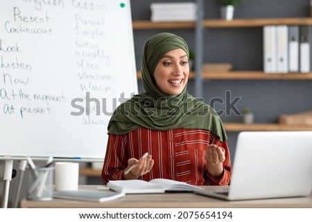 E-learning Concept. Smiling female arabic teacher in headscarf making video call, explaining English grammar rules to students. Young muslim woman talking and looking at pc screen, sitting at desk