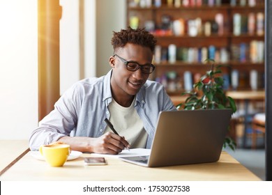 E-learning Concept. Young African American Student With Laptop Taking Notes During Breakfast In Cafe