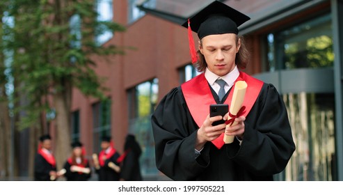 E-learning, Education And People Concept. Happy Smiling Caucasian Male Graduate Student Standing At The Street With Smartphone And Typing Message With Pleasure Smile 