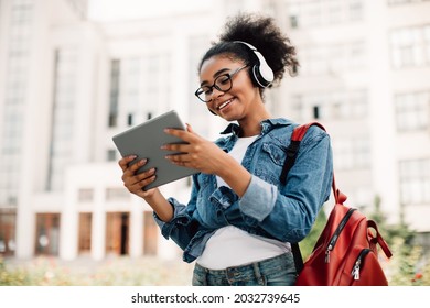 E-Learning. Happy Black Female Student Using Tablet Computer Wearing Headphones Standing Near Modern University Building Outside. Teenager Girl Browsing Internet Outdoors. Modern Online Education