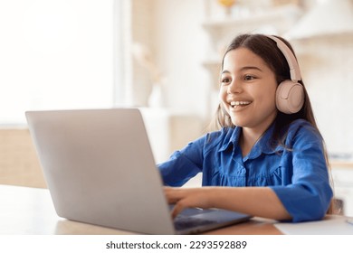 E-Learning. Happy Schoolgirl Studying Online Using Laptop Wearing Headphones At Home, Sitting At Desk. Kid Girl Having Distance Class Typing On Computer And Doing Homework. Modern Remote Education - Powered by Shutterstock