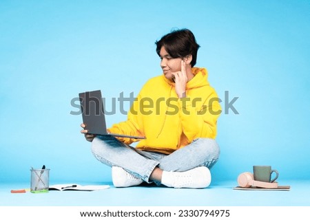 E-Learning Leisure. Japanese Student Guy Using Laptop Computer On Lap, Doing Homework And Making Video Call Online Sitting On Floor Over Blue Studio Background. Internet and Education
