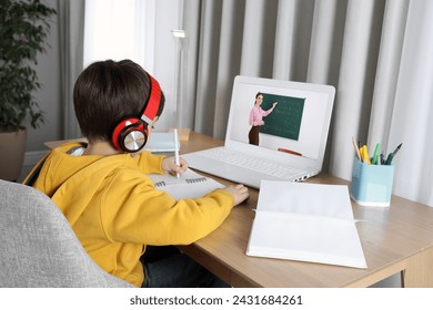 E-learning. Little boy taking notes during online lesson at wooden table indoors