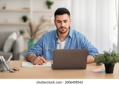 E-learning And Online Training. Portrait Of Focused Jewish Man In Kipa Working Or Studying Remotely At Home Office, Writing In His Notebook With Pen, Taking Notes, Using Laptop Looking At Screen