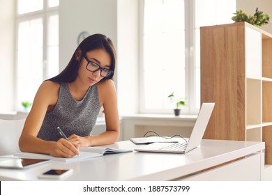 E-learning And Remote Education. Serious Young Girl In Eyeglasses Using Online Educational Tools. Diligent Student Taking Notes While Attending Video Lecture Or Watching Webinar On Laptop Computer