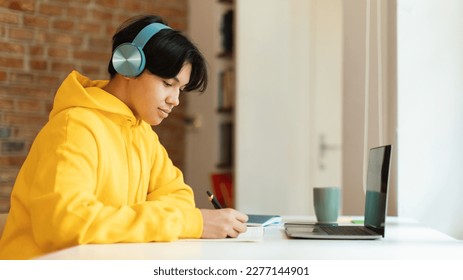 E-Learning. Side View Of Korean Teenager Boy At Laptop Taking Notes Learning Remotely At Home. Student Guy Wearing Headphones Doing His Homework Online On Computer. Panorama - Powered by Shutterstock