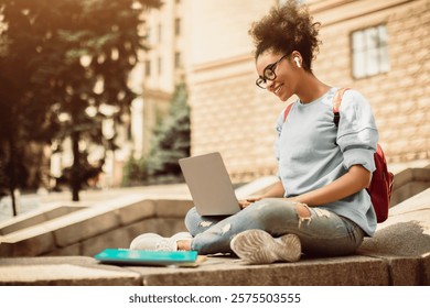 E-Learning. Side-View Of Black Female Student With Laptop Watching Educational Webinar Or Attending Online Lecture Wearing Earbuds And Eyeglasses Sitting Outdoors. Distance College Education