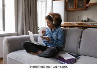 E-learning using modern wireless technology, on-line class with tutor, gain new knowledge. African teenager girl wear headphones listen foreign language audio course, make exercise sit on sofa at home