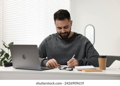 E-learning. Young man taking notes during online lesson at white table indoors