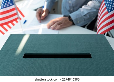 Election In USA. A Male Voter Votes At A Polling Station . The Ballot Box Is In The Foreground With An American Flag