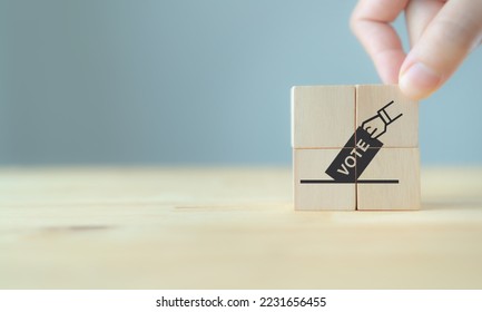Election vote concept. Hand putting paper in the voting box symbol on wooden cube blocks. Political election campaign logo. Applicable as part of badge design. Voting and polling symbols design.  - Shutterstock ID 2231656455