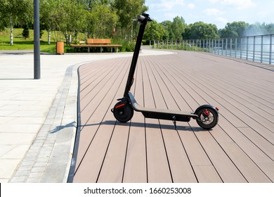 An electric black scooter stands on the bandwagon on the street. City park with wooden flooring along the promenade with railings. Sunny summer day. Modern city transport. - Shutterstock ID 1660253008