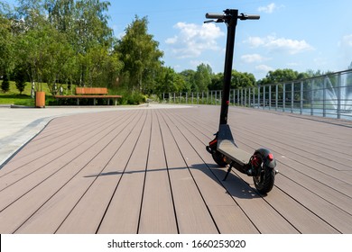 An electric black scooter stands on the bandwagon on the street. City park with wooden flooring along the promenade with railings. Sunny summer day. Modern city transport. - Shutterstock ID 1660253020