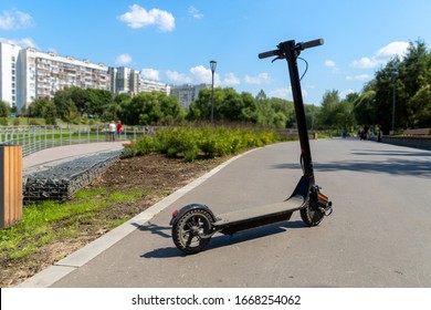 An electric black scooter stands on the bandwagon on the street. City park with wooden flooring along the promenade with railings. Sunny summer day. Modern city transport. - Shutterstock ID 1668254062