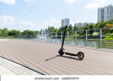 An electric black scooter stands on the bandwagon on the street. City park with wooden flooring along the promenade with railings. Sunny summer day. Modern city transport. - Shutterstock ID 1668254074