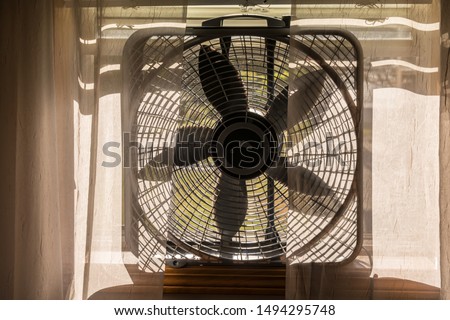 Electric Box Fan Sitting on a Window Sill with Sheer Lace Curtains.