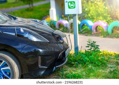 An Electric Car Is Charging In A Parking Space On A City Street Next To The House.Тaken In Backlit And Sun Glare.