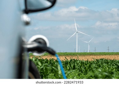 Electric car with EV plug charges from charging station against windfarms on field. Auto stands against distant windmills - Shutterstock ID 2282471933
