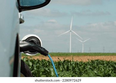 Electric car with EV plug on station with renewable form of energy produced on windfarms. Automobile stands against wind turbines on field - Shutterstock ID 2280972629