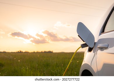 Electric car with opened charging socket cap and charger plugged in, at a public electric charging station near the highway at sunset - Shutterstock ID 2027277722