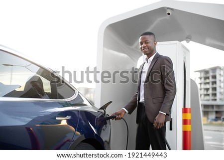 Electric cars, EV concept, eco friendly fuel. Portrait of young smiling black man in business clothes, recharging his modern luxury electric car